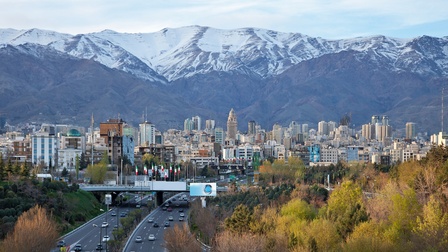 Blick auf die Skyline der Stadt Teheran, gelegen vor dem Elburs-Gebirges mit verschneiten Gipfeln, im Vordergrund sieht man eine befahrene Straße und herbstliche Bäume.