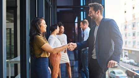 Zwei Personen auf Balkon stehend reichen sich freundlich die Hände, im Hintergrund weitere Personen im Gespräch