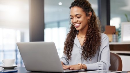Eine Frau sitzt in einem Büro vor einem Laptop an einem Schreibtisch. Die Person tippt auf der Tastatur des Laptops. Neben dem Laptop sind ein Tablet und ein Smartphone
