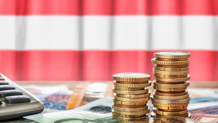 Im Vordergrund sind zwei Münzstapel, unter denen verschiedene Geldscheine liegen. In der linken Bildhälfte ist ein Taschenrechner. Dahinter hängt eine Flagge in den Farben rot-weiß-rot