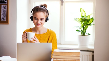 Person mit dunklen geschlossenen Haaren mit gelben Pullover trägt Kopfhörer, hält eine Tasse in den Händen und sitzt bei einem Tisch und blickt gespannt auf einen Laptop, Bücher liegen daneben