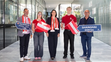 Fünf Personen stehen in einem modernen, verglasten Innenbereich nebeneinander. Zwei Personen halten jeweils ein blaues Schild. Das linke Schild trägt den Text „Team AUSTRIA 2026, 48 Teilnehmer, 42 Berufe, 1 Ziel“. Das rechte Schild zeigt „SHANGHAI 8500 km“. Drei Personen halten rot-weiß-rote Österreich-Flaggen. Mehrere Personen tragen rote Schals mit der Aufschrift „SkillsAustria“