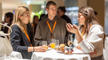 Zu sehen sind Personen, die an einem runden Tisch stehen. Auf diesem sind Gläser und Snacks  platziert. 
