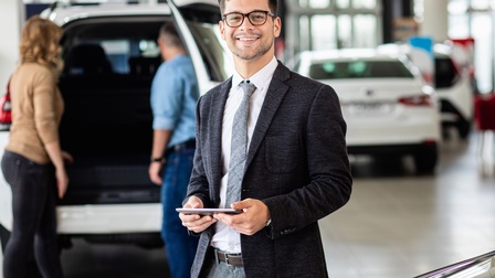 Lächelnde Person mit Tablet in Händen, im Hintergrund weitere Personen und Autos
