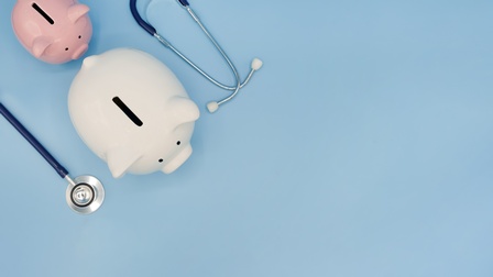 In Vogelperspektive: Auf einem blauen Hintergrund sind in der oberen linke Ecke zwei hintereinander stehende Sparschweine. Das vordere ist weiß und größer als das hintere. Das kleinere hintere ist rosa. Die beiden sind umgeben von einem Stethoskop.