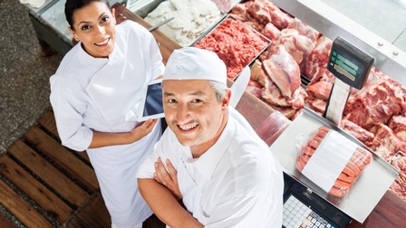Zwei lächelnde Personen in weißem Arbeitsgewand nach oben blickend, daneben Frischetheke mit Fleischwaren