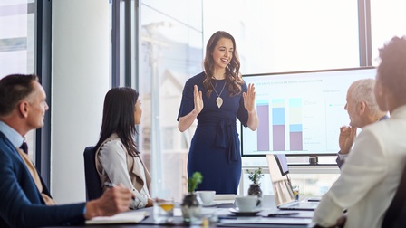 Personen sitzen an Tisch in einem Büroraum mit großen Fenstern, aus denen Tageslicht herein kommt. Eine Person präsentiert, die Präsentation ist auf einem Bildschirm sichtbar und enthält Diagramme. 