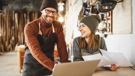 Zwei lächelnde Personen mit Wollmützen in Schürzen stehen vor einem aufgeklappten Notebook in einer Werkstatt.