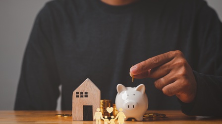 Detailansicht einer Hand, die Münze in Sparschwein wirft, daneben Modell eines Holzhäuschens und Illustration von vier Figuren mit Herze über Kopf