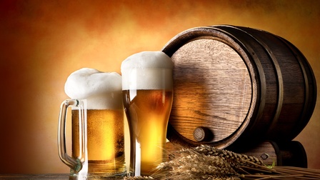 Auf einem Holztisch sind Getreideähren, zwei unterschiedliche Biergläser mit Bier sowie ein kleines Bierfass