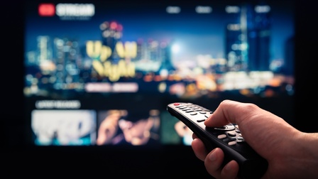 Die rechte Hand einer Person hält eine Fernsehbedienung und deutet damit horizontal auf einen Bildschirm im Hintergrund, auf dem verschwommene Bilder sind