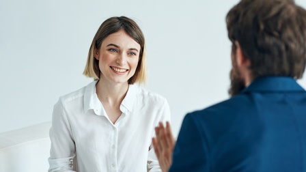 Person mit kurzen Haaren und weißer Bluse sitzt und hält ein Klemmbrett sowie einen Stift und blickt auf eine Person in Unschärfe ihr gegenüber