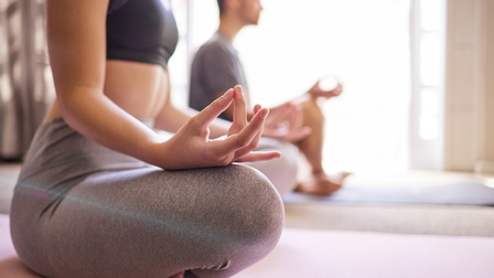 Nahaufnahme von Händen und Schneidersitz während der Meditation, im Hintergrund sitzt eine weitere Person