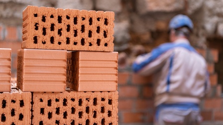 Im Vordergrund sind sechs Ziegelsteine übereinander gestapelt. Im Hintergrund ist der Rücken einer Person mit Helm, die direkt vor einer Mauer aus Ziegeln steht