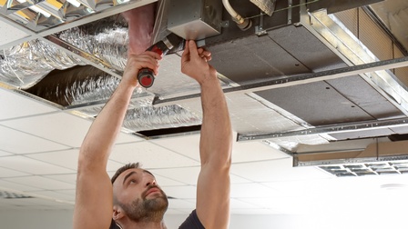 Person mit Bart hält Bohrmaschine Kopfüber an Lüftungsgerät