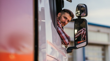 Person mit kurzen Haaren, Bart und kariertem Hemd blickt von der Beifahrerseite eines LKWs aus dem Fenster zurück