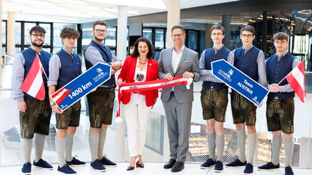 Porträt von acht Personen, die nebeneinander stehen. Die drei Personen links und rechts tragen jeweils eine Lederhose und halten ein Schild, auf dem 1400 Kilometer und Euroskills steht. Die beiden Personen in der Mitte tragen jeweils ein Sakko und halten einen Banner in rot-weiß-rot, auf dem Skills Austria steht
