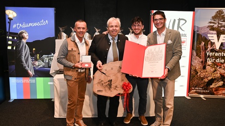Vier Personen stehen bei einer Ehrung vor Werbebannern, halten eine Urkunde, eine Medaille und eine große Holz-Uhr als Auszeichnung in die Kamera.