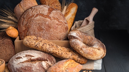 Auf einem dunklen Holzuntergrund liegen verschiedene Backwaren in einer Kiste und davor, zum Beispiel Brot, Baguette oder Weckerl. In der Kiste sind außerdem die oberen Teile einer Getreidepflanze. Vor den Backwaren verstreut liegen Körner.
