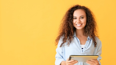 Eine Person mit lockigem, langem dunkelbraunem Haar steht vor gelbem Hintergrund. Sie hält mit beiden Händen ein Tablet