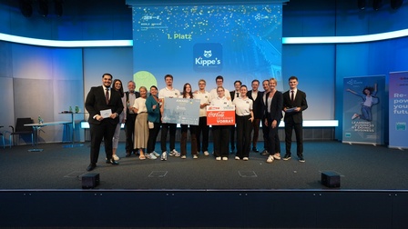 Gruppenfoto auf einer Veranstaltungsbühne. Mehrere Personen stehen vor einer großen Projektionsfläche. In der Projektion stehen die Texte „1. Platz“ und „Kippe's“. Einige Personen halten eine Auszeichnungstafel sowie einen Gutschein