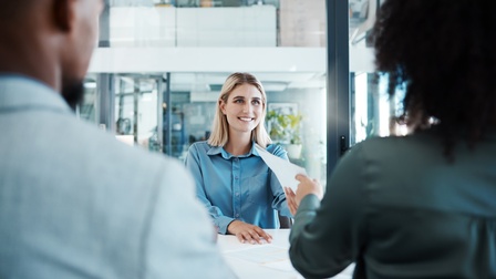 Ein an einem Tisch mit anderen Personen sitzende Person bekommt  Dokumente ausgehändigt.