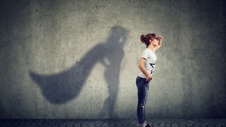 Person mit langen dunklen geschlossenen Haaren mit Brille steht mit aufrechter Haltung und blickt in die Ferne während an der Wand ein Schatten der Person mit einem Superhero-Cape erscheint