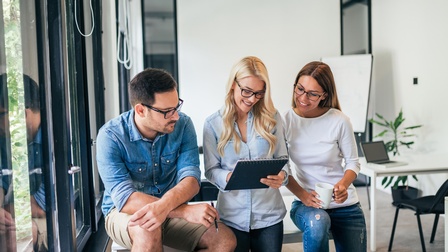 Drei Personen stehen nebeneinander. Die mittlere Person hält einen Notizblock, auf den alle blicken. Im Hintergrund ist ein Whiteboard
