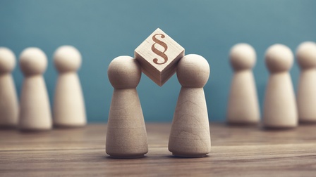 Auf einer hölzernen Oberfläche stehen im Vordergrund zwei kleine Holzfiguren, zwischen deren Köpfen ein Holzwürfel gehalten wird, auf dem ein Paragrafensymbol ist. Im Hintergrund stehen weitere Holzfiguren