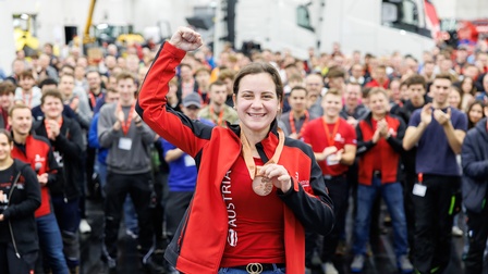 Porträt einer Person, die eine Medaille um den Hals hat und die rechte Faust in die Luft hält. Hinter der Person stehen sehr viele Personen, die applaudieren