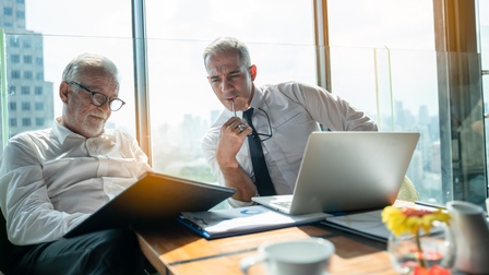 Zwei Personen sitzen an Arbeitstisch und blicken auf Clipboard, das eine Person am Schoß hält, andere Person hält Brille an Mund, am Tisch aufgeklapptes Notebook und Unterlagen, im Hintergrund Fenster