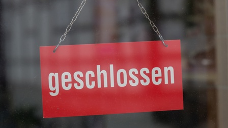 Vor einem grauen, verschwommenen Hintergrund sieht man ein Schild in der Bildmitte, das an einer Metallkette hängt. Das Schild ist rot und trägt die Inschrift 