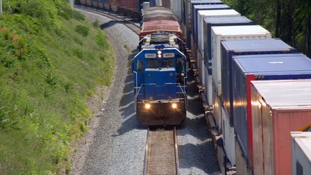 Auf Gleisen fährt ein langer Zug mit vielen Containern. Neben dem Zug fährt auf den anderen Gleisen eine kurze Lok mit ein paar Containern. Links und rechts von den Gleisen sind Bäume und ein grüner Hügel