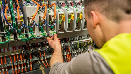 Rückenansicht einer Person, die sich nach vorne beugt und ihre linke Hand auf einem grauen, sehr kleinen Kästchen hat, auf dem ein grüner Zettel klebt. Um dieses Kästchen sind viele weitere sowie viele unterschiedliche Kabel.