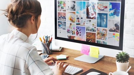 Rückenansicht einer Person, die vor einem Bildschirm sitzt, auf dem Bilder zu sehen sind. Sie tippt in eine Tastatur, die auf einem Schreibtisch aus Holz platziert ist, neben Stiften, Post-its, einer Topffplanze und einer Tasse mit Kaffee. 