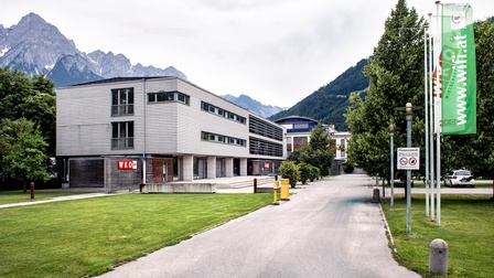 Moderner Gebäudekomplex der Wirtschaftskammer Tirol in alpiner Umgebung: Ein mehrgeschossiger, kubischer Bau mit Holzfassade und großen Fensterflächen steht links entlang einer Zufahrtsstraße. Rechts säumen Bäume und Fahnen mit WIFI-Branding den Weg. Im Hintergrund erheben sich bewaldete Hügel und schroffe Berggipfel. Die Szene wirkt ruhig, gepflegt und gut erreichbar.