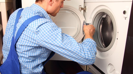 Person in blauer Arbeitskleidung schraubt am Bullauge einer Waschmaschine