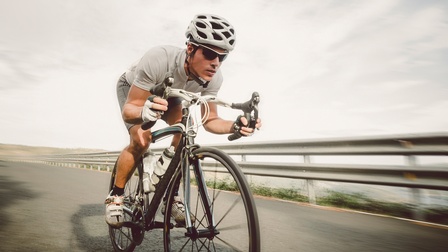 Person in Sportkleidung, Fahrradhelm, Sportsonnenbrille, fingerlosen Handschuhen sitzt auf einem Fahrrad und fährt damit eine betonierte Straße hinunter