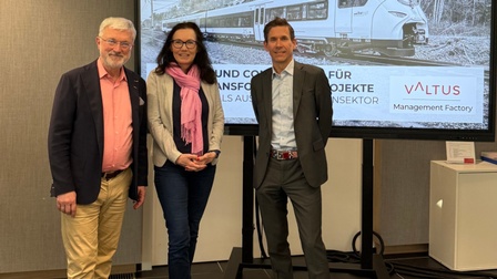 Drei Personen stehen vor einem Bildschirm während einer Veranstaltung.