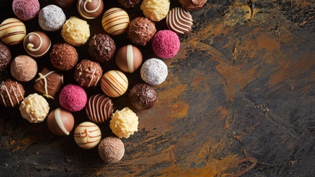 Topshot vieler, kleiner, runder Pralinen, die auf einer dunklen Oberfläche liegen. Manche Pralinen sind dunkel, andere hell, andere rosa. Manche haben Striche oben, manche sind uneben