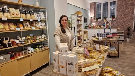 Porträt einer Person mit dunklen Haaren, Brille und hellen Oberteilen, die hinter einem Tisch steht. Auf dem Tisch sind verschiedene, verpackte Lebensmittel. Hinter der Person ist ein Regal, in dem weitere Lebensmittel sind