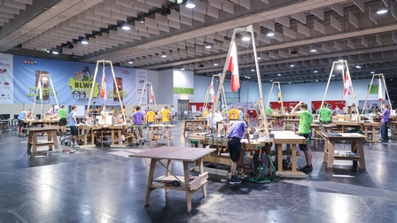 In einer großen Halle sind zahlreiche Arbeitsstationen mit Werkbänken angeordnet. An den Stationen arbeiten oder stehen mehrere Personen. Über den Arbeitsbereichen befinden sich hohe Gestelle mit Kennzeichnungen und Fahnen