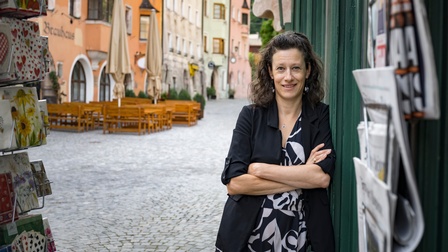 Eine Person steht mit verschränkten Armen an einer Hauswand und blickt in die Kamera. Im Hintergrund sind eine gepflasterte Straße, bunte Gebäude und Sitzgelegenheiten im Freien zu sehen.