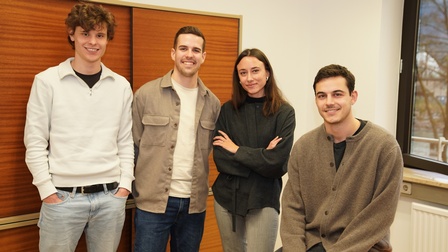Vier junge Erwachsene stehen bzw. sitzen in einem Büroraum und lächeln in die Kamera. Drei Personen stehen nebeneinander, eine Person sitzt rechts auf einer Tischkante. Der Raum hat weiße Wände, ein großes Fenster und Holzschränke im Hintergrund.
