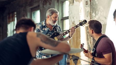 Drei Personen beim Musizieren: Eine Person spielt auf abgewinkeltes Bein gestützt Gitarre und singt, eine weitere Person danebensitzend spielt Gitarre, im Vordergrund verschwommen Person an Schlagzeug sitzend