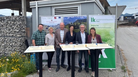 Sechs Personen stehen vor Informationsbannern des Regionalmanagements und der Landwirtschaftskammer Tirol bei einer Projektvorstellung im Freien. Die Gruppe posiert vor einem Gewerbegebäude, auf Stehtischen sind Namensschilder der Beteiligten aufgestellt. Im Hintergrund sind ein Gewerbegebiet sowie Berglandschaft zu sehen.