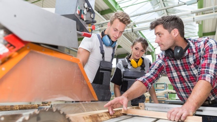 Lehrlinge (ein Mann, eine Frau) schauen einem Ausbildner zu, wir er Holz mit einer Kreissäge bearbeitet