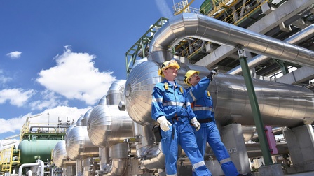 Zwei Personen in Schutzausrüstung mit gelben Helmen gehen entlang Raffinerietanks, im Hintergrund blauer Himmel mit Wolken