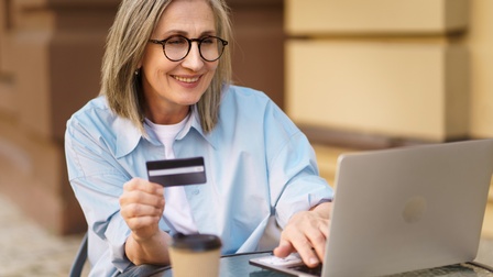 Lächelnde Person mit Brillen Bistrotisch vor aufgeklappten Laptop sitzend hält Bankkarte in Händen und blickt auf Monitor, auf Tisch verschwommen im Vordergrund Kaffeebecher