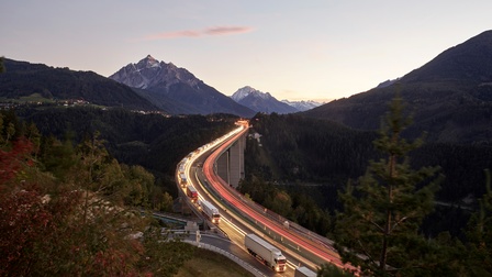 Landschaft in Dämmerlicht mit Autobahn mit Gefährten, eine Fahrbahn in Bewegungsunschärfe, die andere befahren von Lastkraftwagen, ringsum bewaldete und beschneite Berge